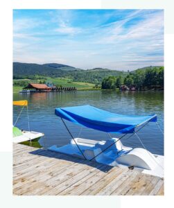 A dock with paddle boats, one with a blue canopy, on a calm lake surrounded by green hills and distant buildings under a partly cloudy sky.